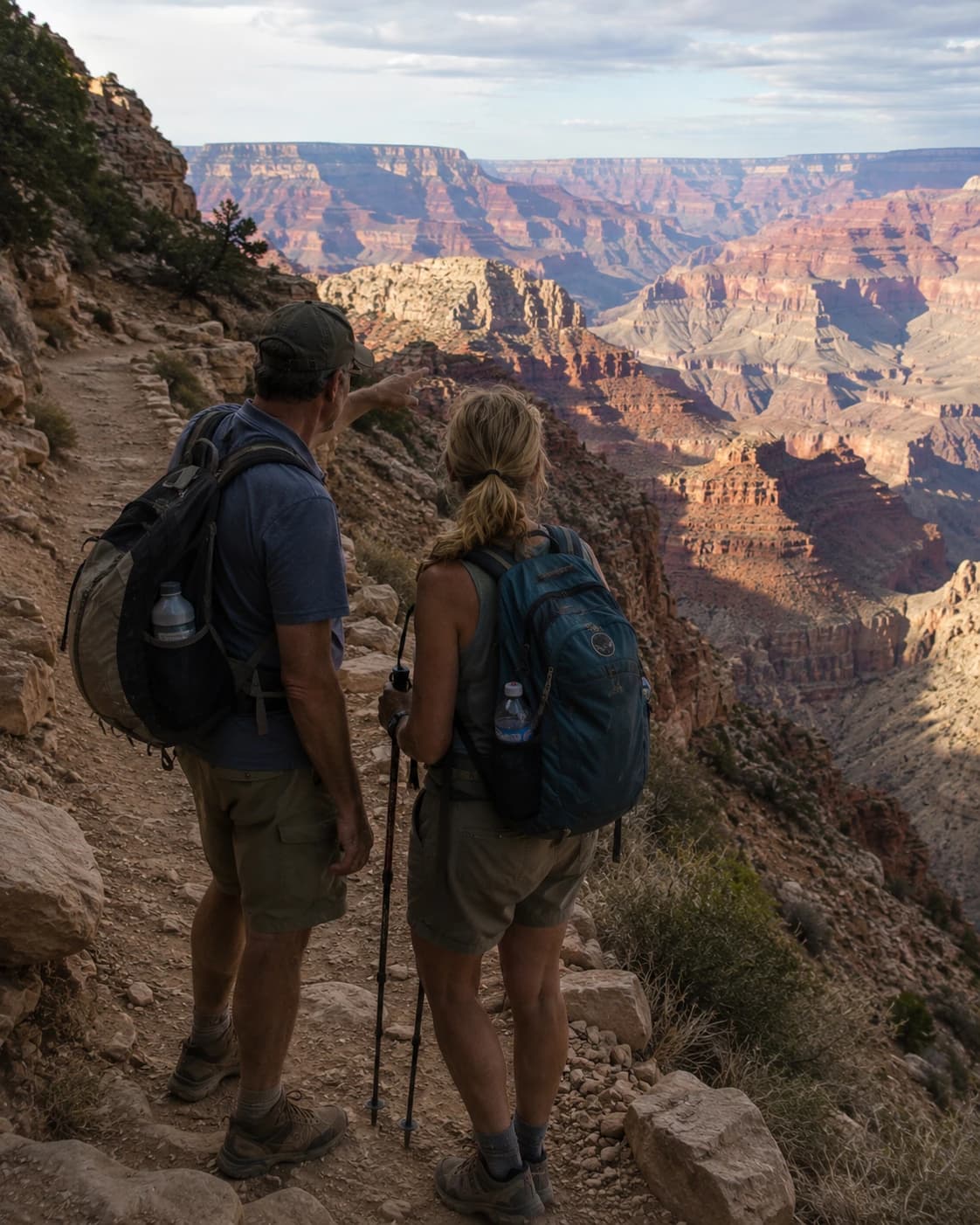 A couple with backpacks pausing on a trail overlook at the Grand Canyon, looking out over the canyon at golden hour